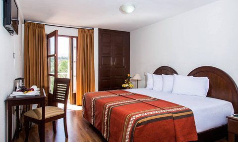 Ein helles Hotelzimmer mit einem großen Bett, einem Tisch und Stühlen, sowie einem Fenster mit Vorhängen, das Tageslicht hereinlässt.