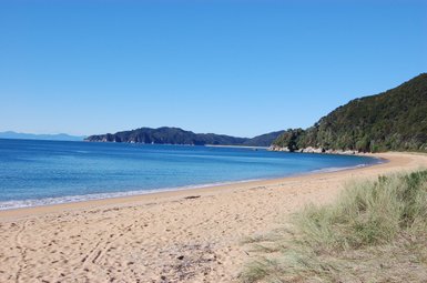 Ein ruhiger Strand mit feinem Sand, umgeben von sanften Hügeln und klarem, blauem Wasser unter einem strahlend blauen Himmel.