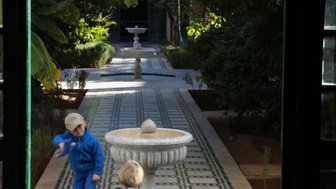 Zwei Kinder spielen fröhlich in einem Garten mit einem Brunnen, umgeben von üppigem Grün und bunten Fliesen.