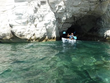 Ein Kajakfahrer paddelt durch klares Wasser in eine Höhle, umgeben von weißen Felsen und strahlendem Sonnenlicht.