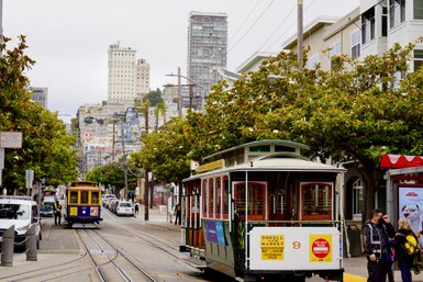 Eine Straßenbahn fährt durch eine belebte Straße in San Francisco, umgeben von hohen Gebäuden und üppigem Grün.