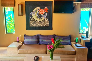 Ein gemütliches Sofa mit gestreiften Kissen steht vor einem bunten Wandteppich und einem Fernseher in einem hellen Raum.