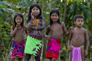 Vier Kinder in traditioneller Kleidung, die mit kulturellen Tätowierungen geschmückt sind, stehen lächelnd in der Natur.