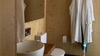 Ein modernes Badezimmer mit Holzverkleidung, einem Waschbecken und zwei weißen Bademänteln, die an der Wand hängen.