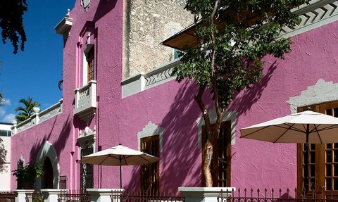 Eine lebhafte, pinkfarbene Fassade eines Gebäudes mit großen Fenstern und Sonnenschirmen im Freien.