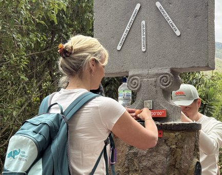 Eine Frau mit einem Rucksack interagiert mit einem Steinsäule, während ein Mann sie mit einem Smartphone filmt.