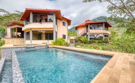Zwei moderne Häuser mit roten Dächern stehen neben einem glitzernden Pool, umgeben von üppigem Grün und Bergen im Hintergrund.