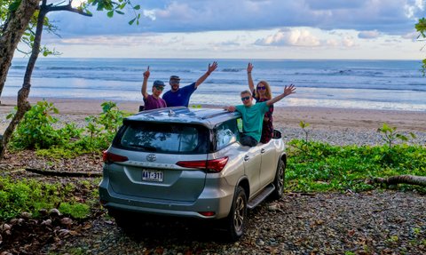 Eine Gruppe von vier Personen steht fröhlich neben einem silbernen Auto am Strand von Costa Rica, umgeben von üppigem Grün.