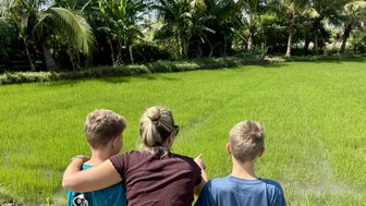 Eine Frau und zwei Jungen stehen am Rand eines Reisfeldes und betrachten die grüne Landschaft unter einem blauen Himmel.