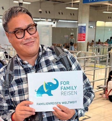 Ein lächelnder Reiseleiter hält ein Schild mit der Aufschrift 'FOR FAMILY REISEN' am Flughafen in einer belebten Umgebung.