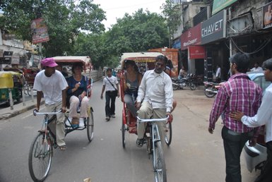 Zwei Rikschafahrer transportieren Touristen auf einer belebten Straße in Delhi, umgeben von Geschäften und Bäumen.