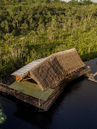 Eine malerische Lodge mit Strohdach steht am Ufer eines ruhigen Gewässers, umgeben von üppigem, grünem Dschungel.