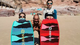 Drei Personen stehen am Strand, halten bunte Bodyboards und lächeln fröhlich in die Kamera, umgeben von einer schönen Landschaft.