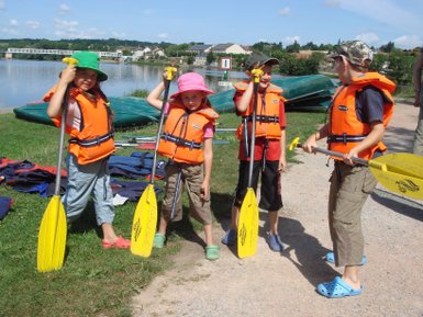 Vier Kinder in Schwimmwesten stehen am Ufer eines Gewässers, bereit zum Kanufahren, mit Paddeln in der Hand.
