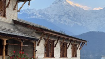 Ein traditionelles nepalesisches Lodge-Gebäude mit Holzverzierungen und blühenden Blumen im Vordergrund, umgeben von majestätischen Bergen.