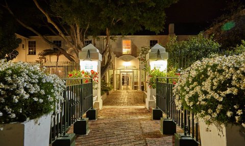 Der Eingangsbereich des Windsor Hotels in Hermanus ist nachts beleuchtet, umgeben von blühenden Blumen und grünen Bäumen.
