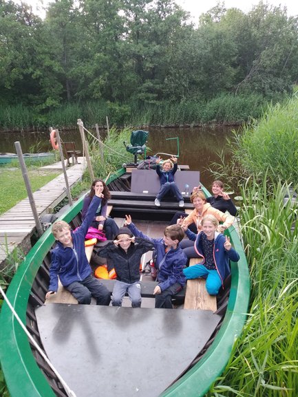 Eine Gruppe von Kindern und Erwachsenen sitzt fröhlich in einem Boot, umgeben von üppigem Grün und einem ruhigen Gewässer.