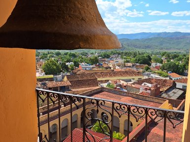 Eine große Glocke hängt über einem Balkon mit Blick auf eine malerische Stadtlandschaft und grüne Hügel im Hintergrund.