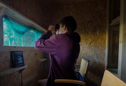 Ein junger Mann beobachtet mit einem Fernglas durch ein Fenster in einer Holzhütte, umgeben von Natur.