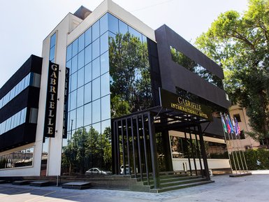 Das moderne Gabrielle International Hotel mit großen Glasfenstern spiegelt die umliegenden Bäume und den blauen Himmel wider.