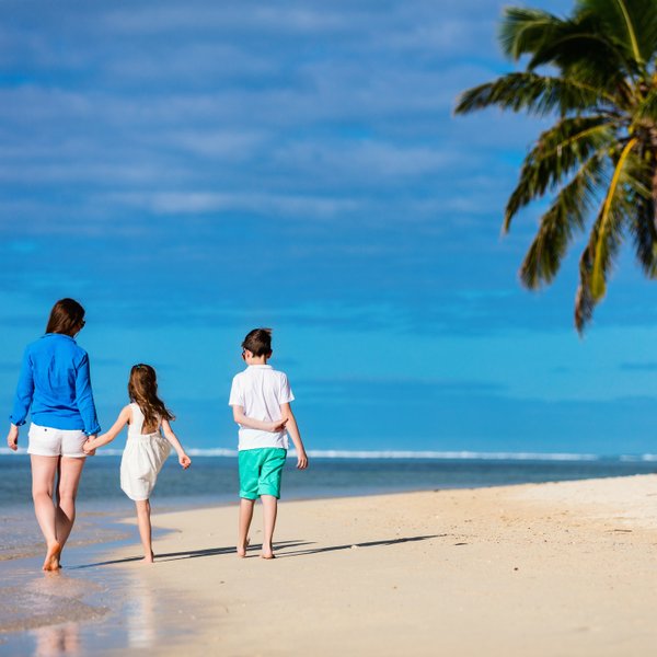 Eine Mutter und zwei Kinder spazieren barfuß am Strand entlang, umgeben von Palmen und einem strahlend blauen Himmel.