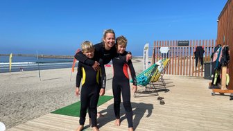 Eine Frau umarmt zwei Kinder in Neoprenanzügen am Strand, bereit zum Surfen, mit Wellen im Hintergrund.