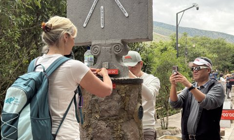 Eine Frau mit einem Rucksack interagiert mit einem Steinsäule, während ein Mann sie mit einem Smartphone filmt.