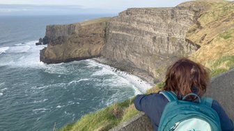 Eine Person mit einem Rucksack steht an der Kante der Klippen und blickt auf die raue See und die zerklüfteten Felsen.