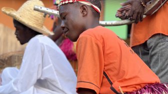 Ein junger Tänzer in einem orangefarbenen Shirt und einem bunten Rock spielt mit einem Stock auf einem Holzblock.