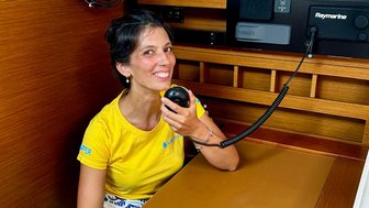 Eine fröhliche Frau mit einem Mikrofon sitzt in einem Boot und lächelt, umgeben von Holzverkleidung und modernen Geräten.