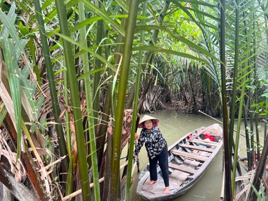 Eine Frau in einem traditionellen Hut steht auf einem Boot, umgeben von üppigem Grün im Mekong-Delta.