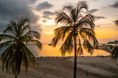 Palmen rahmen den Strand bei Sonnenuntergang ein, während Menschen am Wasser entspannen und ein Boot in der Ferne fährt.