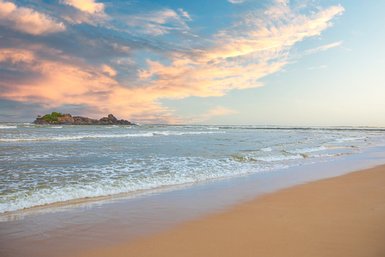 Ein ruhiger Strand mit sanften Wellen, die an den goldenen Sand schlagen, unter einem farbenfrohen Himmel bei Sonnenuntergang.