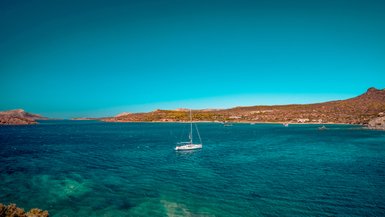 Ein Segelboot gleitet sanft über das glitzernde, türkisfarbene Wasser, umgeben von sanften Hügeln und klarem Himmel.