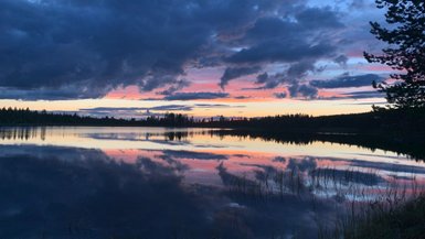 Ein ruhiger See spiegelt die dramatischen Wolken und den farbenfrohen Sonnenuntergang wider, umgeben von dichten Wäldern.