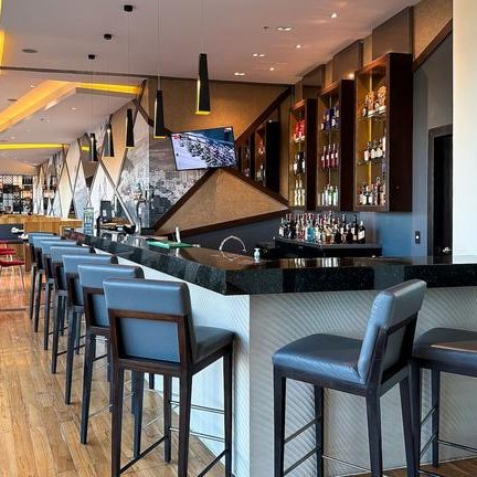 Eine elegante Bar mit modernen Stühlen und einer gut gefüllten Spirituosenregal im Hintergrund, beleuchtet von warmem Licht.