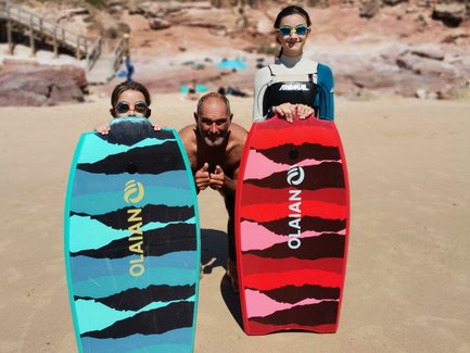 Drei Personen stehen am Strand, halten bunte Bodyboards und lächeln fröhlich in die Kamera, umgeben von einer schönen Landschaft.