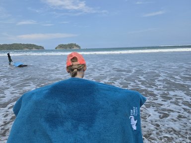 Ein Kind steht barfuß am Strand, umgeben von Wellen und trägt ein großes, blaues Handtuch über den Schultern.