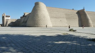 Die massive Zitadelle von Buchara erhebt sich majestätisch unter einem klaren blauen Himmel, umgeben von einem gepflasterten Platz.