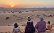 Eine Familie sitzt auf einer Sanddüne und beobachtet den Sonnenuntergang über der Wüste, umgeben von sanften Hügeln.