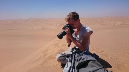 Ein Fotograf sitzt in der Wüste und fokussiert seine Kamera auf die weite, sandige Landschaft unter einem klaren blauen Himmel.