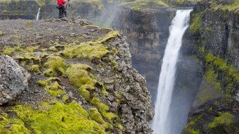 Zwei Fotografen in roten Jacken stehen am Rand einer Klippe und bewundern den majestätischen Haifoss-Wasserfall.