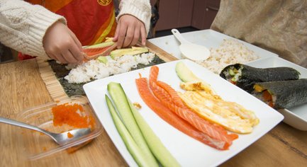 Ein Kind rollt Sushi mit frischem Lachs, Avocado und Gurke auf einem Bambusmatte, während eine Schüssel Reis daneben steht.