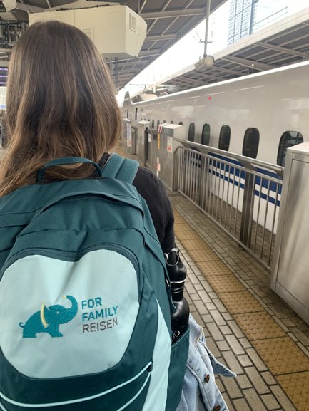Eine Person mit langen Haaren steht an einem Bahnhof, während ein Hochgeschwindigkeitszug im Hintergrund wartet.