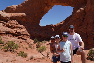 Eine Familie posiert fröhlich vor dem beeindruckenden Felsenbogen im Arches Nationalpark, umgeben von rotem Sandstein.