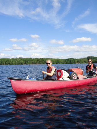 Zwei Personen paddeln in einem roten Kanu auf einem ruhigen See, umgeben von grünen Bäumen und blauem Himmel.