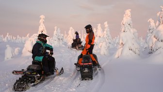 Zwei Schneemobilfahrer in winterlicher Landschaft, umgeben von schneebedeckten Bäumen und sanften Hügeln im Hintergrund.