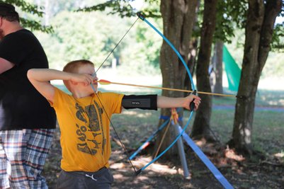 Ein Junge mit einem gelben T-Shirt zielt mit einem Bogen auf ein Ziel, während ein Erwachsener im Hintergrund steht.