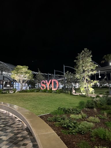 Die leuchtenden Buchstaben 'SYD' stehen prominent in einem stilvollen Gartenbereich bei Nacht.