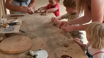 Ein Mann zeigt Kindern, wie man mit Ton arbeitet, während sie an einem Tisch in einem hellen Raum sitzen.